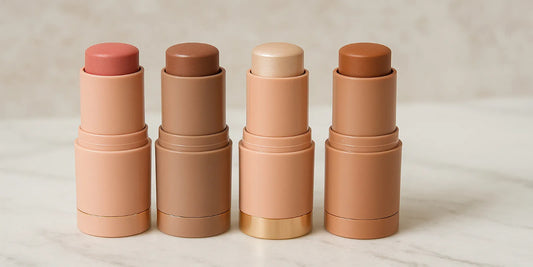 Four makeup sticks—blush, contour, highlighter, and bronzer—arranged on a marble surface with soft lighting, showcasing peach-toned matte tubes.