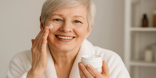 Smiling senior woman in a white bathrobe applying face cream, holding a jar of moisturizer in a bright bathroom with natural warm lighting.