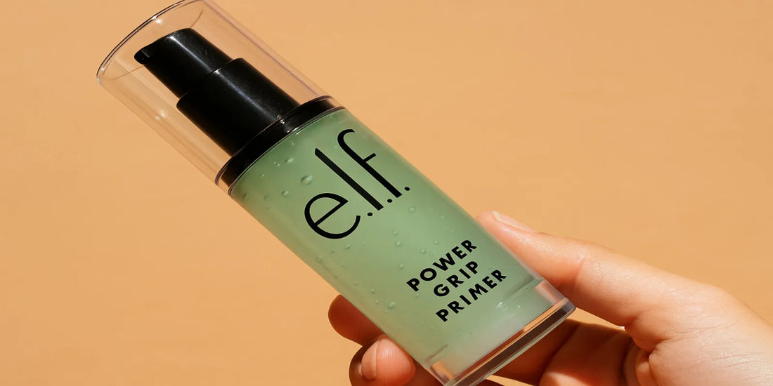 A hand holding a bottle of e.l.f. Power Grip Primer with a clear green gel inside, against a warm peach background, showcasing the product design.
